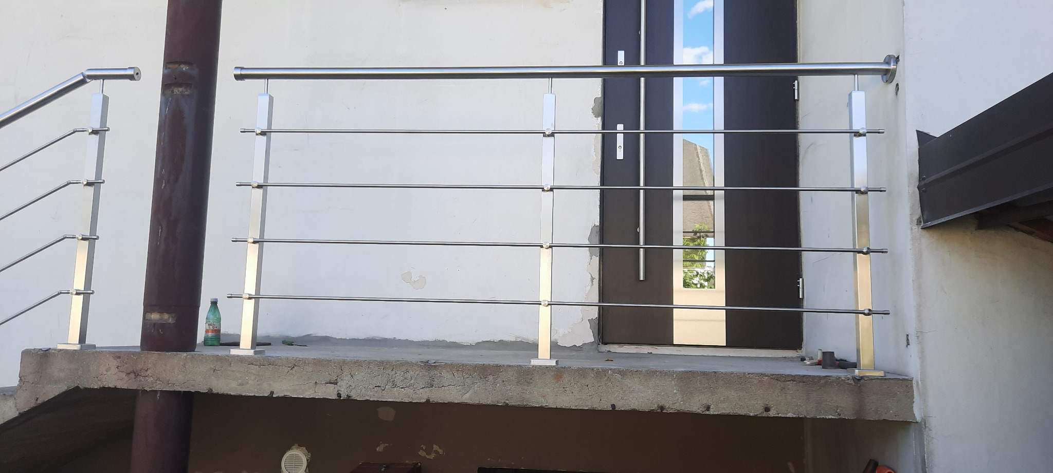 Nowoczesna balustrada ze stali nierdzewnej z poziomymi prętami, zamontowana przy wejściu do budynku z ciemnymi drzwiami z szybami odbijającymi niebo.