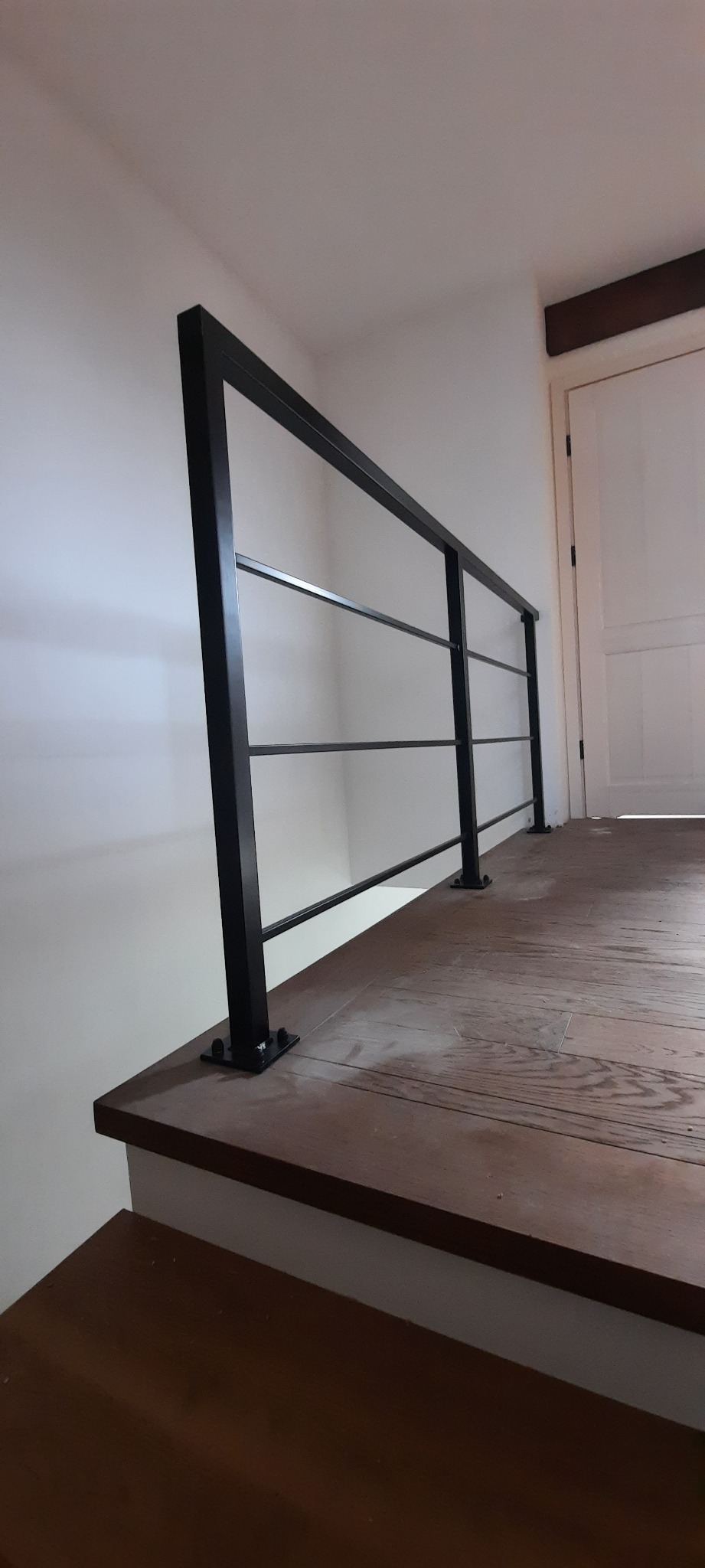 Minimalistyczna, czarna poręcz z poziomymi prętami na drewnianym podeście wewnątrz budynku, widoczne mocowania do podłoża.