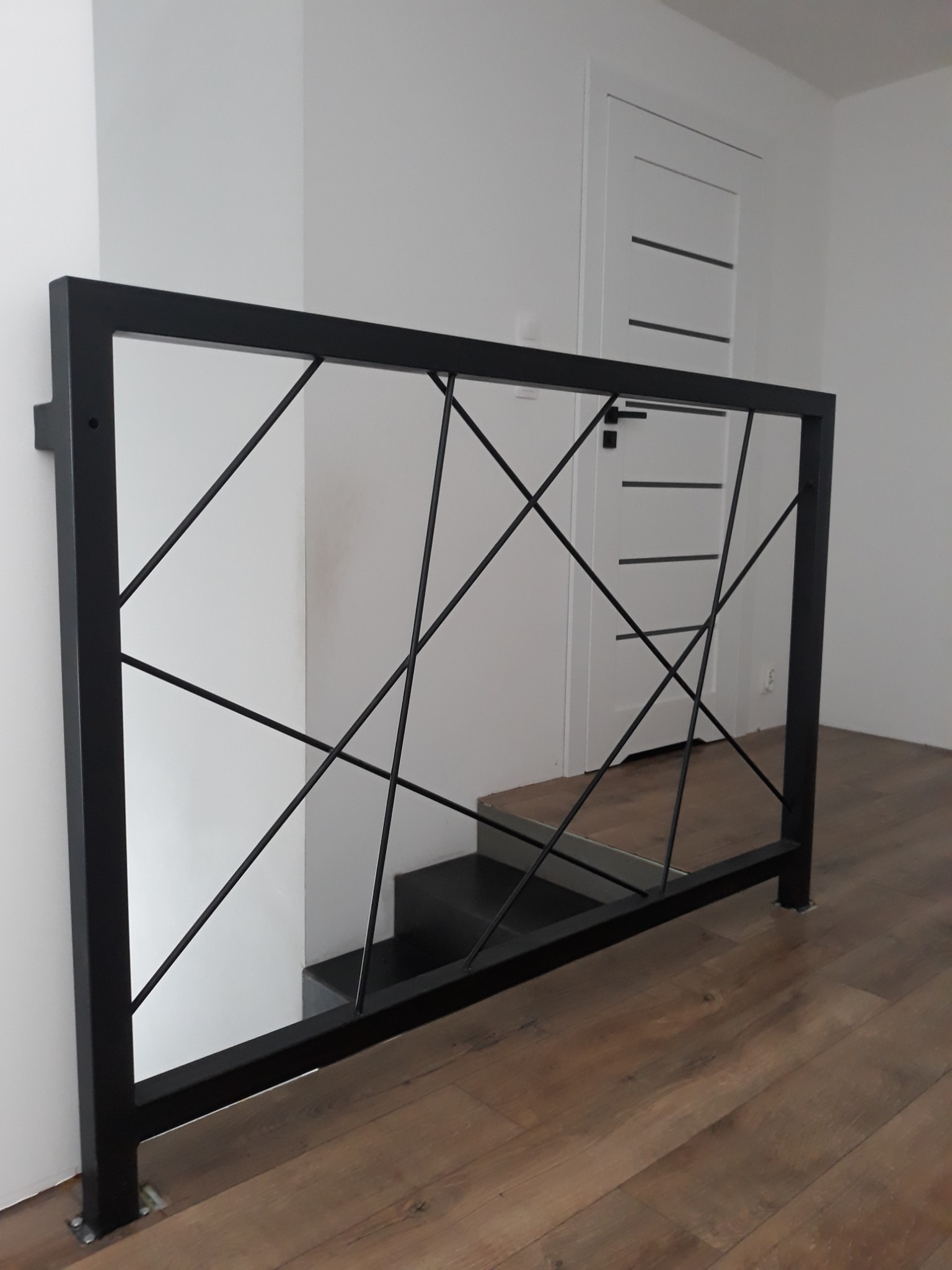 Czarna balustrada wewnętrzna z geometrycznym wzorem z cienkich prętów, widok na fragment schodów i białe drzwi z poziomymi wgłębieniami.