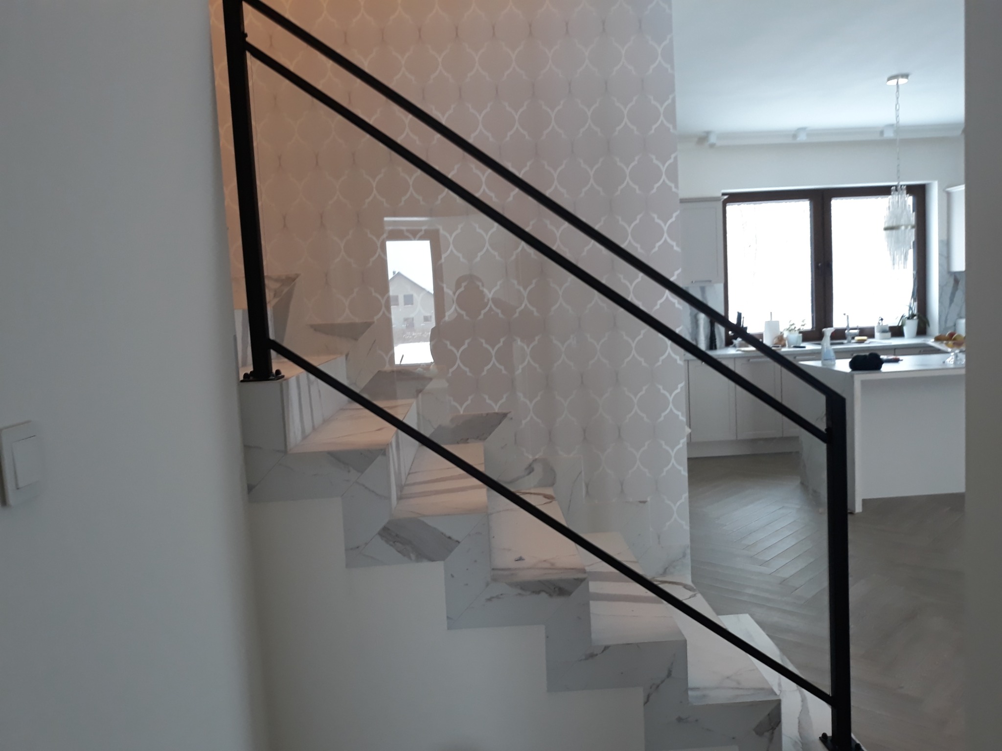 Schody wykończone marmurem z czarną, minimalistyczną poręczą na tle wzorzystej tapety i widoku na kuchnię.