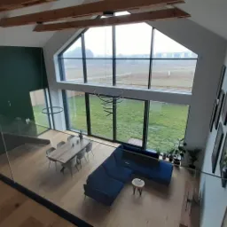 Wysoki salon z antresoli, widok na drewniane belki stropowe, szklaną balustradę, dużą ścianę okienną z widokiem na zewnątrz, ciemnozieloną ścianę, drewnianą podłogę i niebieski narożnik.