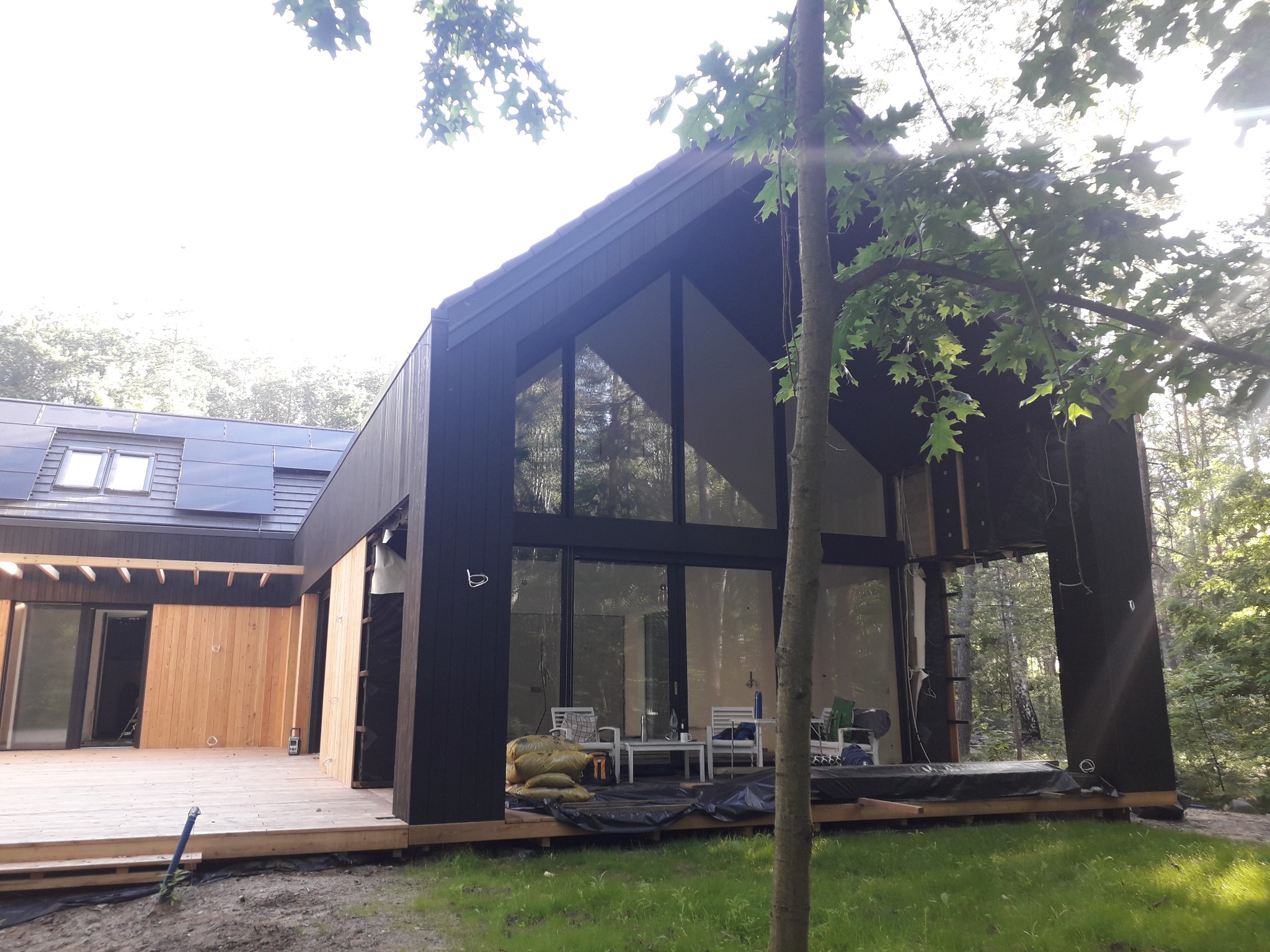 Nowoczesny dom z dużymi przeszkleniami i drewnianą elewacją w trakcie budowy, widoczne worki z materiałami budowlanymi na tarasie.