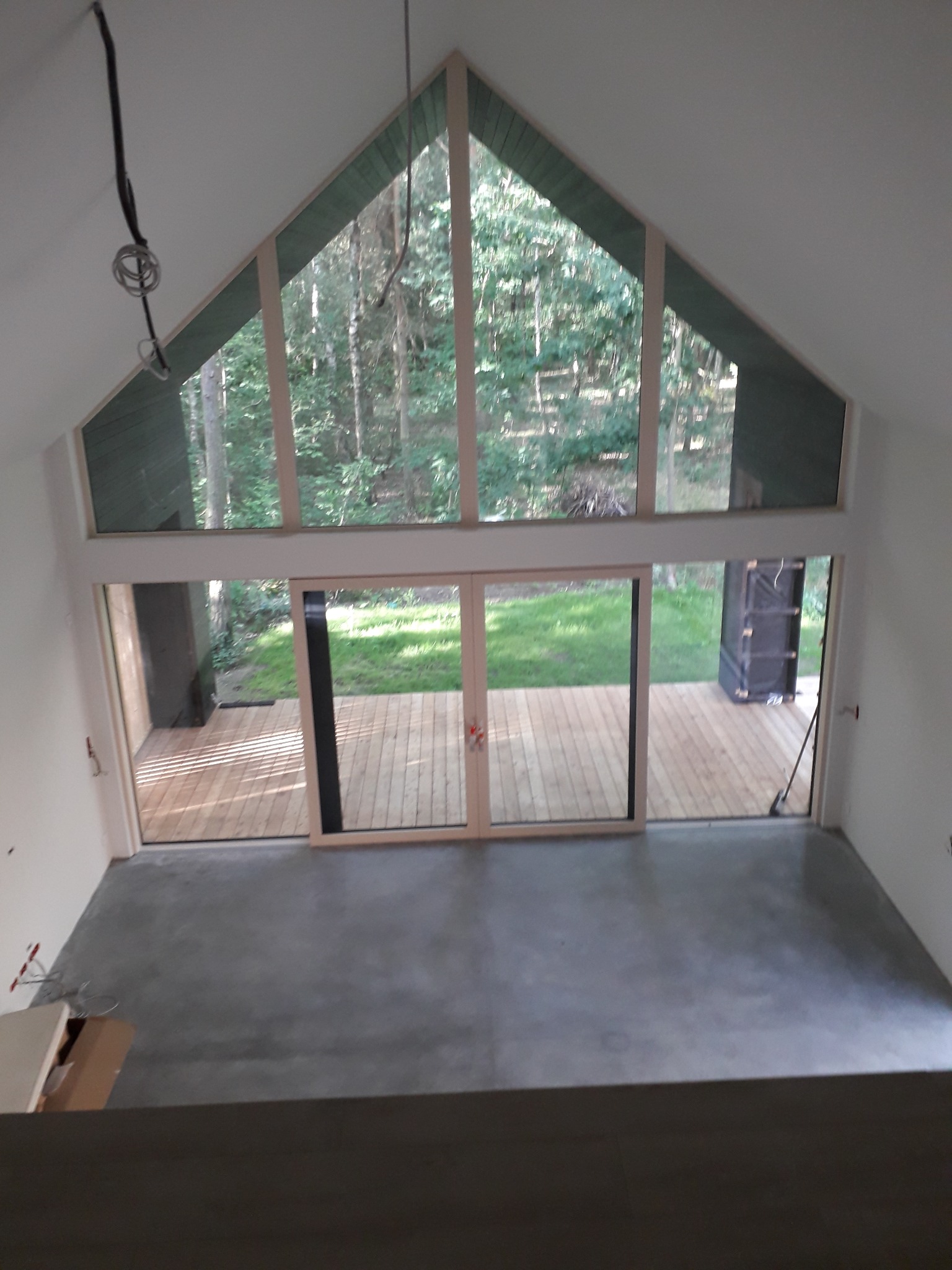 Wysokie okno z widokiem na las w surowym wnętrzu z betonową posadzką i drewnianym tarasem na zewnątrz, widoczne przewody elektryczne na suficie.