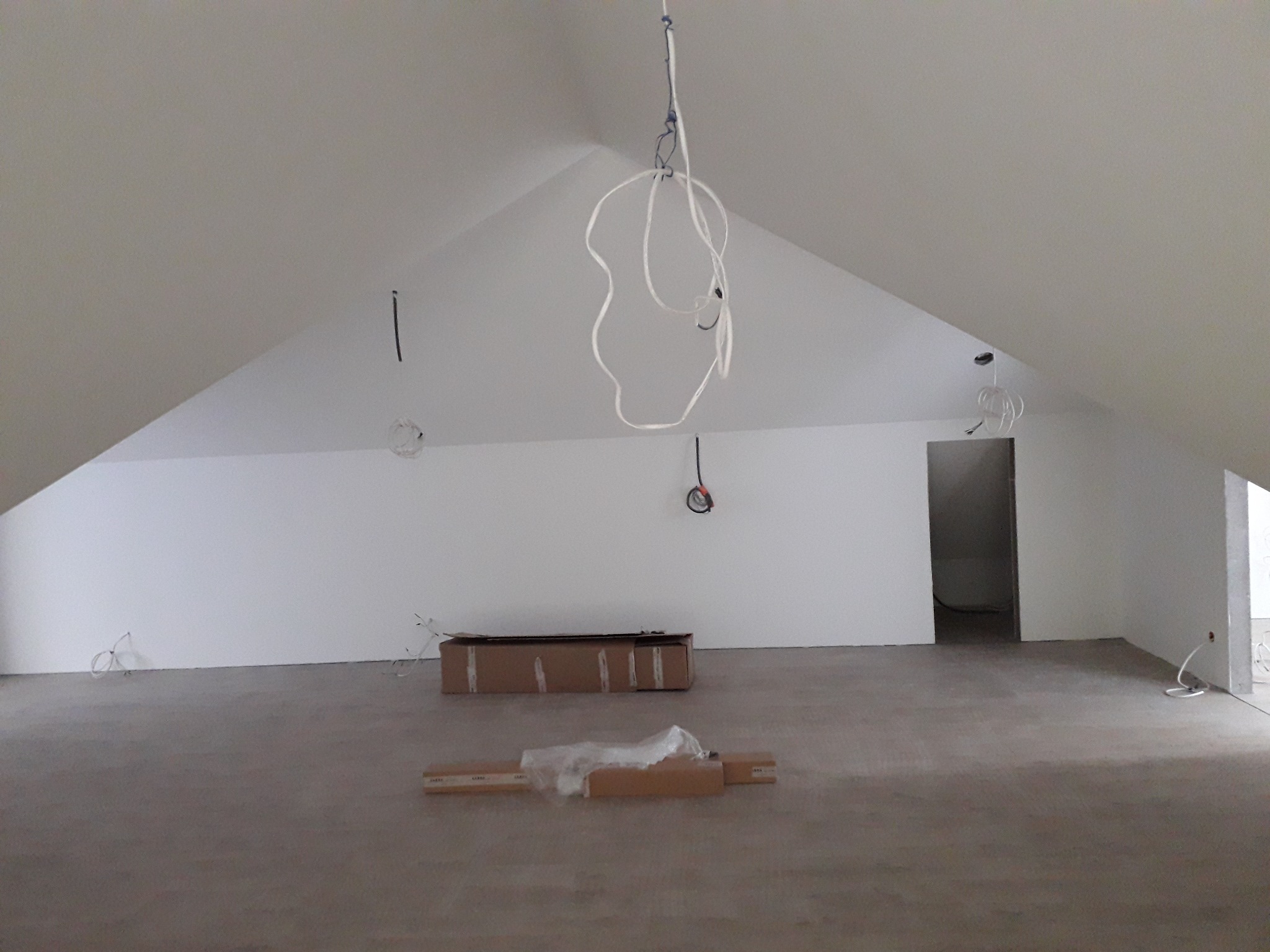 Poddasze w trakcie przygotowania do wykończenia, widoczne przewody elektryczne zwisające z sufitu, surowe ściany i podłoga, oraz materiały budowlane w kartonach.
