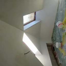 Wnętrze w trakcie prac wykończeniowych, widoczne okno dachowe, ściany pokryte świeżą warstwą gładzi, podłoga zabezpieczona folią malarską, puszki z farbą i narzędzia malarskie.