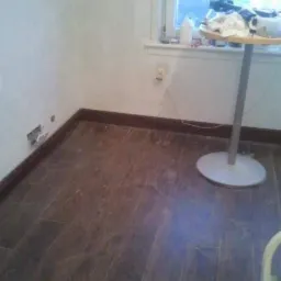 Pokój w trakcie prac wykończeniowych, z ułożoną ciemną podłogą z paneli, widoczne listwy przypodłogowe, ściany przygotowane do malowania z widocznymi gniazdkami i stolik pomocniczy z akcesoriami...