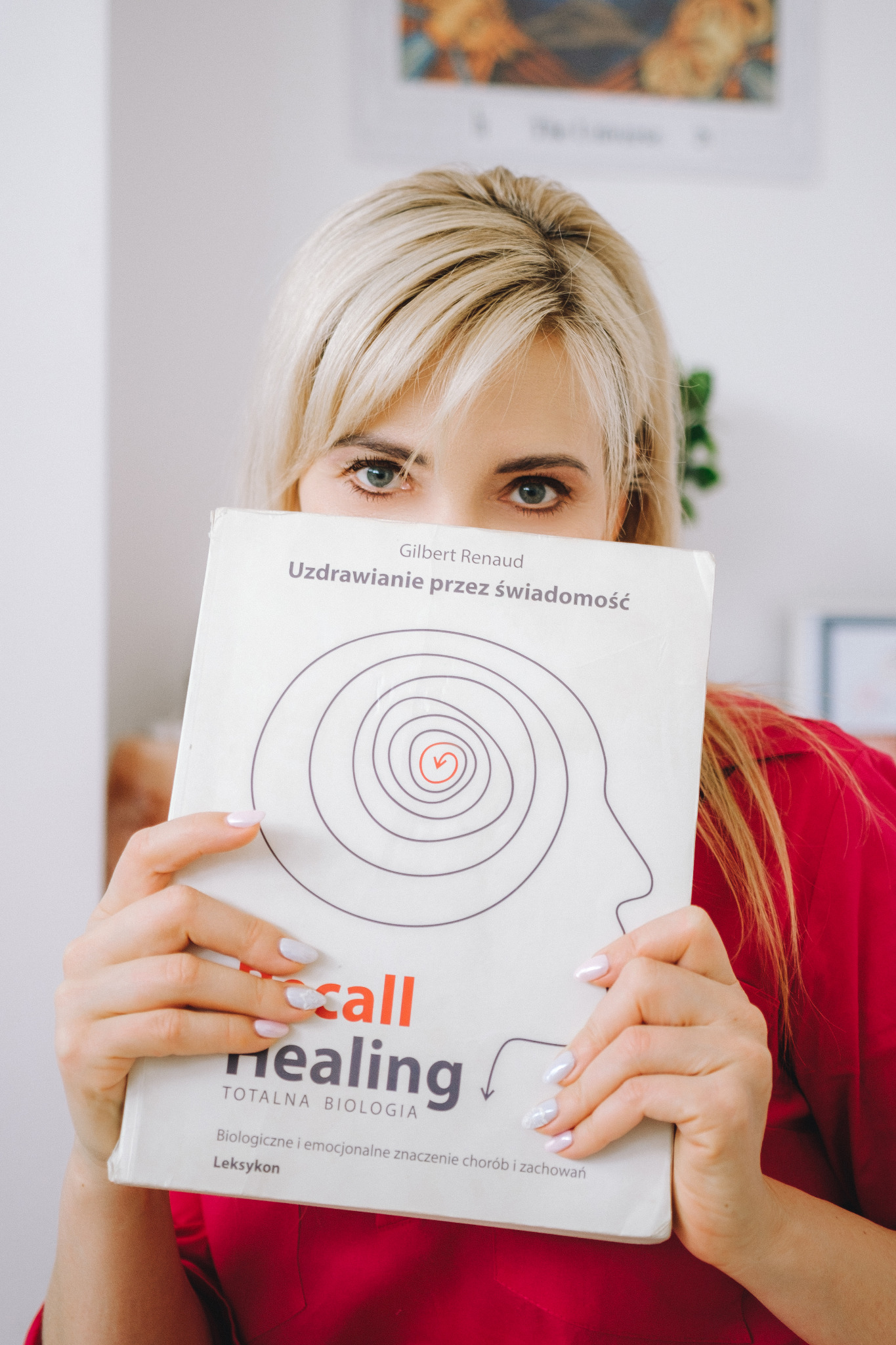 Kobieta z blond włosami zasłania twarz książką 'Uzdrawianie przez świadomość' Gilberta Renauda, z grafiką głowy i spiralą w środku. Widoczne zielone oczy i fragment czerwonej bluzki.