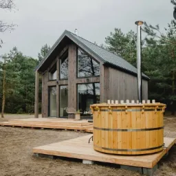 Nowoczesny dom z drewna z dużą wanną z hydromasażem na zewnątrz, otoczony lasem iglastym i drewnianym tarasem.