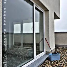 Okno przesuwne na tarasie pokrytym kamykami, wiadro z mopem do mycia okien stoi obok, widoczne odbicie wnętrza pokoju.