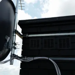 Antena satelitarna i antena naziemna zamontowane na kominie z czarnej cegły, widoczne kable i elementy mocujące na tle pochmurnego nieba.