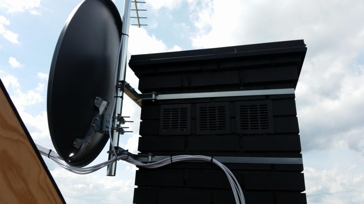 Antena satelitarna i antena naziemna zamontowane na kominie z czarnej cegły, widoczne kable i elementy mocujące na tle pochmurnego nieba.