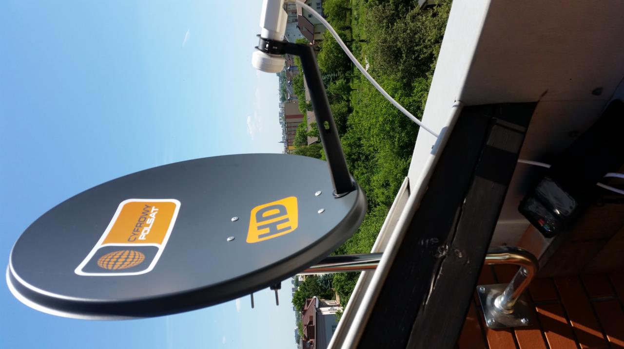 Antena satelitarna Cyfrowy Polsat HD zamontowana na balkonie z widokiem na zieleń miejską i budynki w tle.