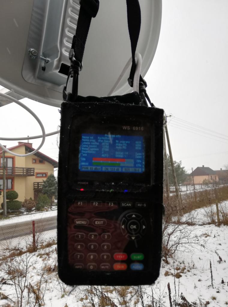 Miernik sygnału satelitarnego WS-6916 zawieszony na czaszy anteny satelitarnej, widoczny ekran z parametrami sygnału na tle zaśnieżonego krajobrazu z budynkami.