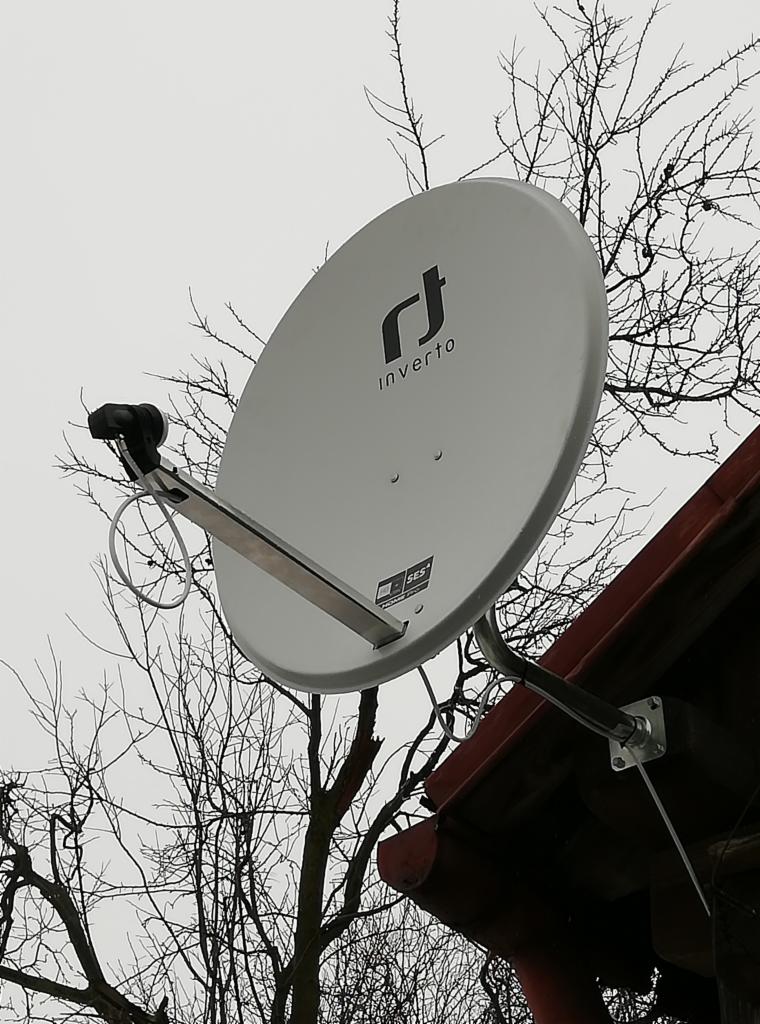 Szara antena satelitarna marki Inverto, zamontowana na dachu z czerwonej dachówki, na tle nagich gałęzi drzew i pochmurnego nieba.