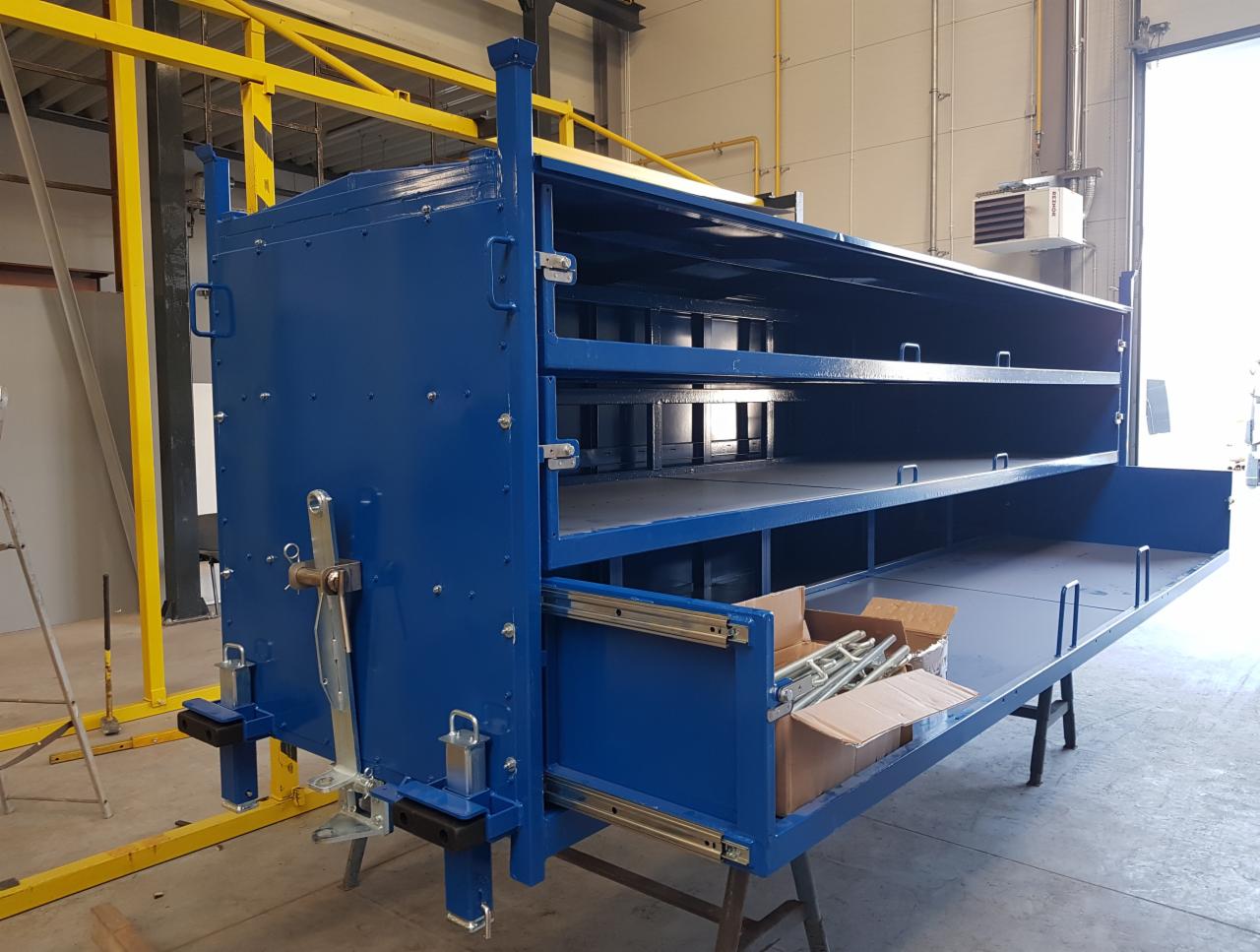 Niebieska metalowa konstrukcja z wysuwanymi półkami, widoczne prowadnice i elementy montażowe w kartonach, ustawiona w hali produkcyjnej z żółtą konstrukcją wsporczą.