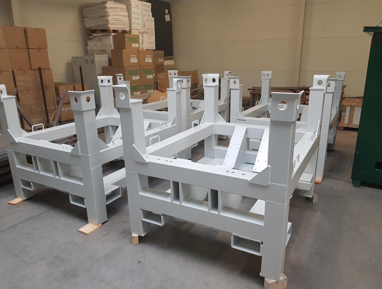 Stalowe ramy konstrukcyjne, pomalowane na szaro, ustawione w hali produkcyjnej na drewnianych podkładkach.