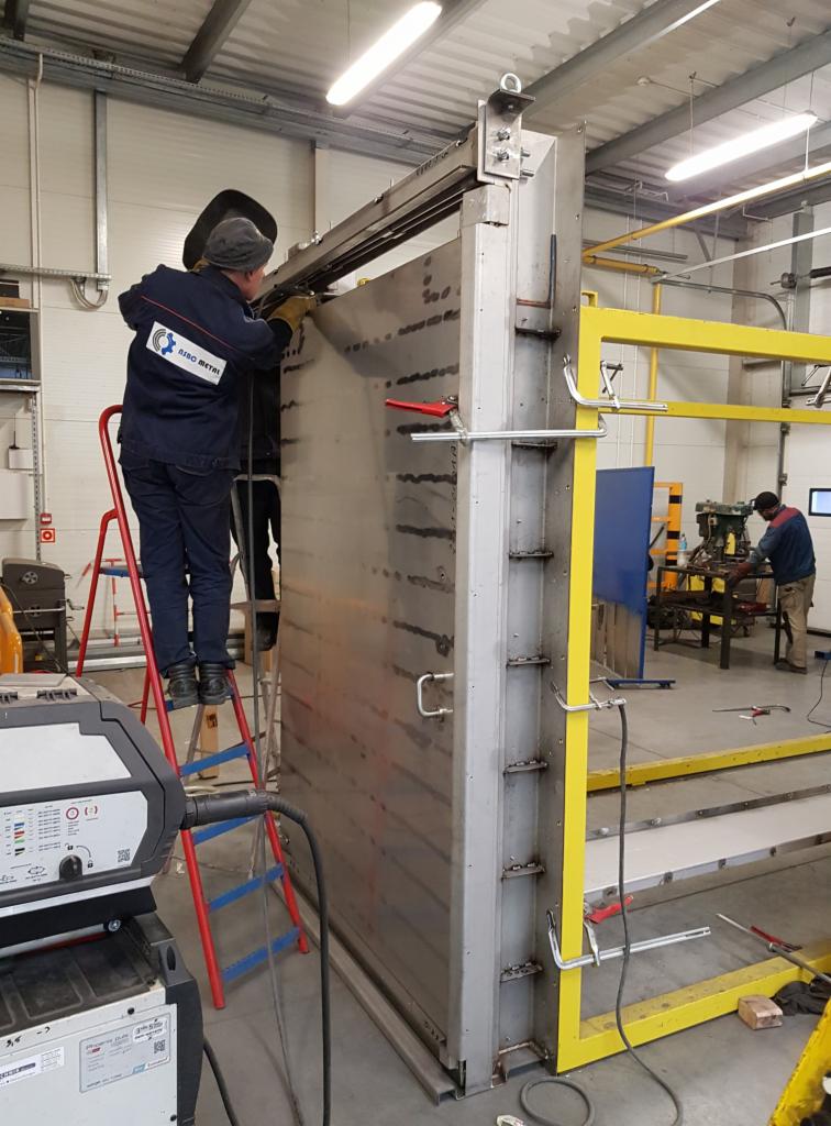 Pracownik w kombinezonie firmy RISO METAL spawa metalową konstrukcję na hali produkcyjnej, stojąc na czerwonej drabinie, widoczny spawacz w masce i spawarka, w tle drugi pracownik przy obróbce metalu.