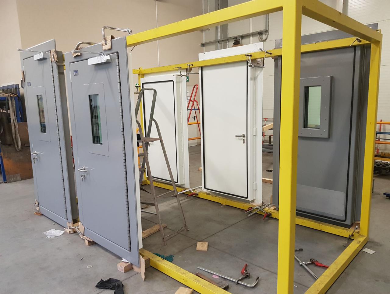Cztery metalowe drzwi z małymi okienkami, częściowo szare i białe, ustawione w żółtej metalowej ramie, prawdopodobnie przed montażem lub transportem.
