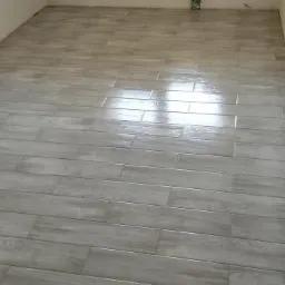 Świeżo ułożona, błyszcząca podłoga z płytek imitujących drewno, ułożonych w jodełkę, z widocznym odbiciem światła i fragmentem ściany z gniazdkiem elektrycznym.