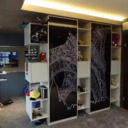 Pokój młodzieżowy z zabudową meblową z motywem wieży Eiffla na frontach, otwarte półki wypełnione różnymi przedmiotami, w tle szara ściana i telewizor.