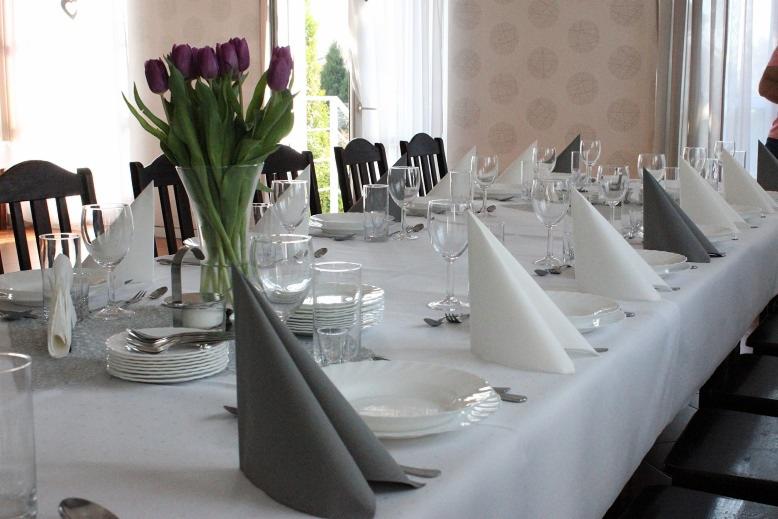 Długi stół nakryty białym obrusem, z szarymi i białymi serwetami ułożonymi w trójkąty, szklankami, talerzami i sztućcami, w wazonie fioletowe tulipany, widok na ogród w tle.