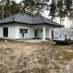 Pracownik na drabinie przy oknie nowo wybudowanego, jednopiętrowego domu z białą elewacją, ciemną dachówką i czarnymi ramami okiennymi, na tle lasu i z zaparkowanym busem firmy budowlanej z reklamą...