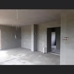 Surowe wnętrze w trakcie prac wykończeniowych: ściany i sufit pokryte szarą warstwą, widoczne wystające przewody elektryczne, puszki instalacyjne i rurka wentylacyjna na podłodze.