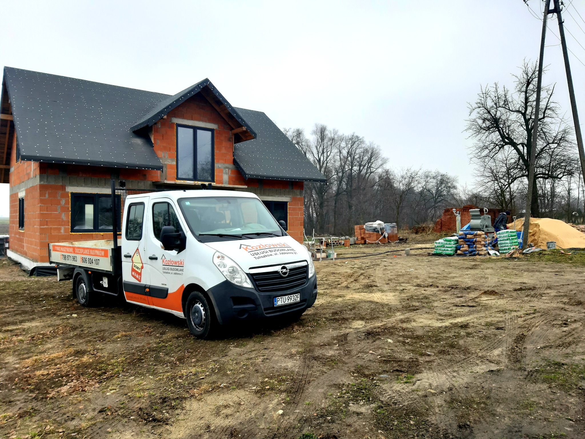 Surowy budynek z czerwonej cegły w trakcie budowy, z oknami i czarnym dachem, na tle szarego nieba i drzew bez liści. Na pierwszym planie biały samochód dostawczy z logo firmy budowlanej i numerami...