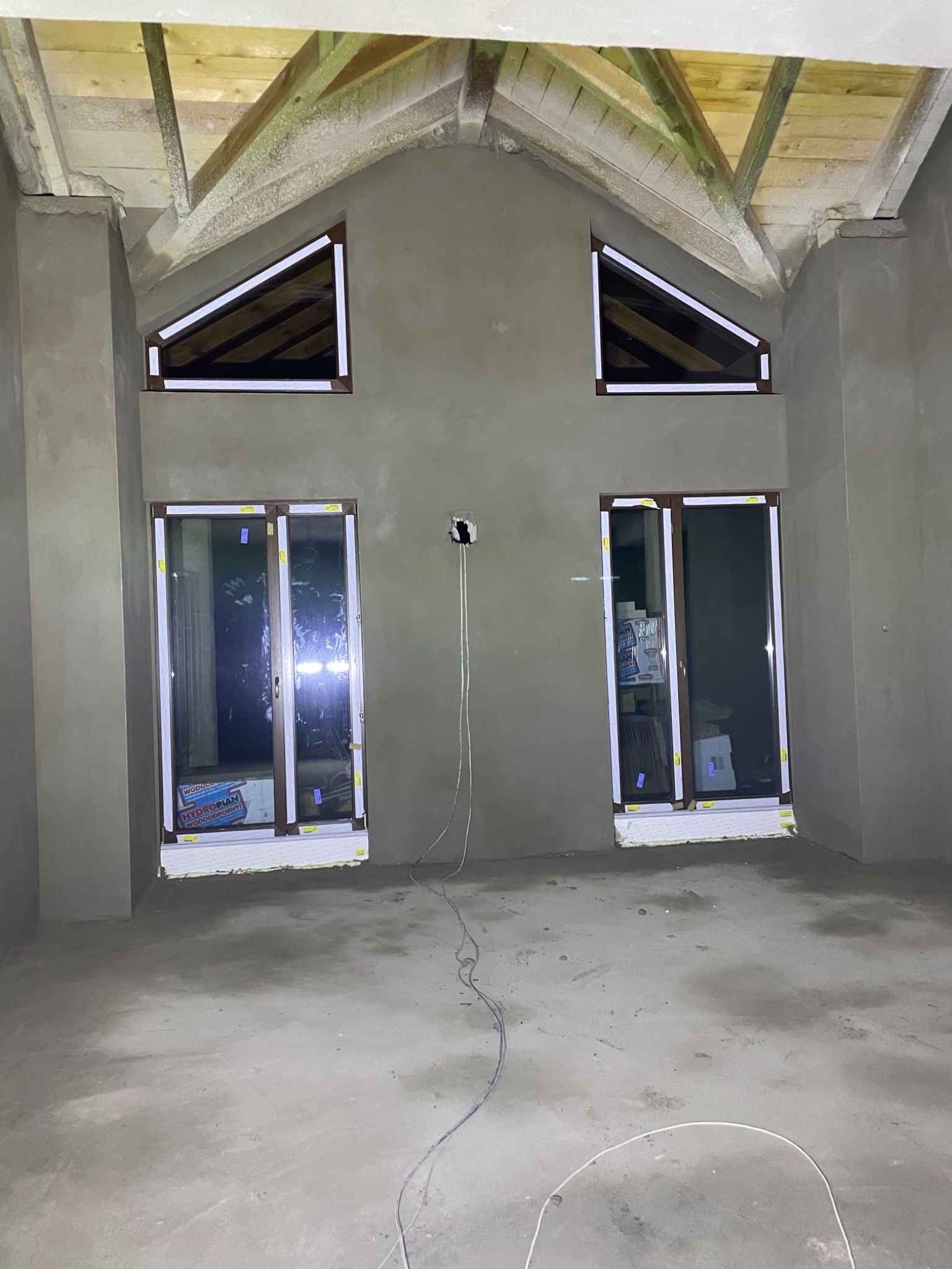 Surowe wnętrze poddasza z drewnianą konstrukcją dachu, świeżo pokryte szarym tynkiem, widoczne dwa okna połaciowe i dwa okna pionowe z brązowymi ramami, zabezpieczone taśmą malarską, wystające...