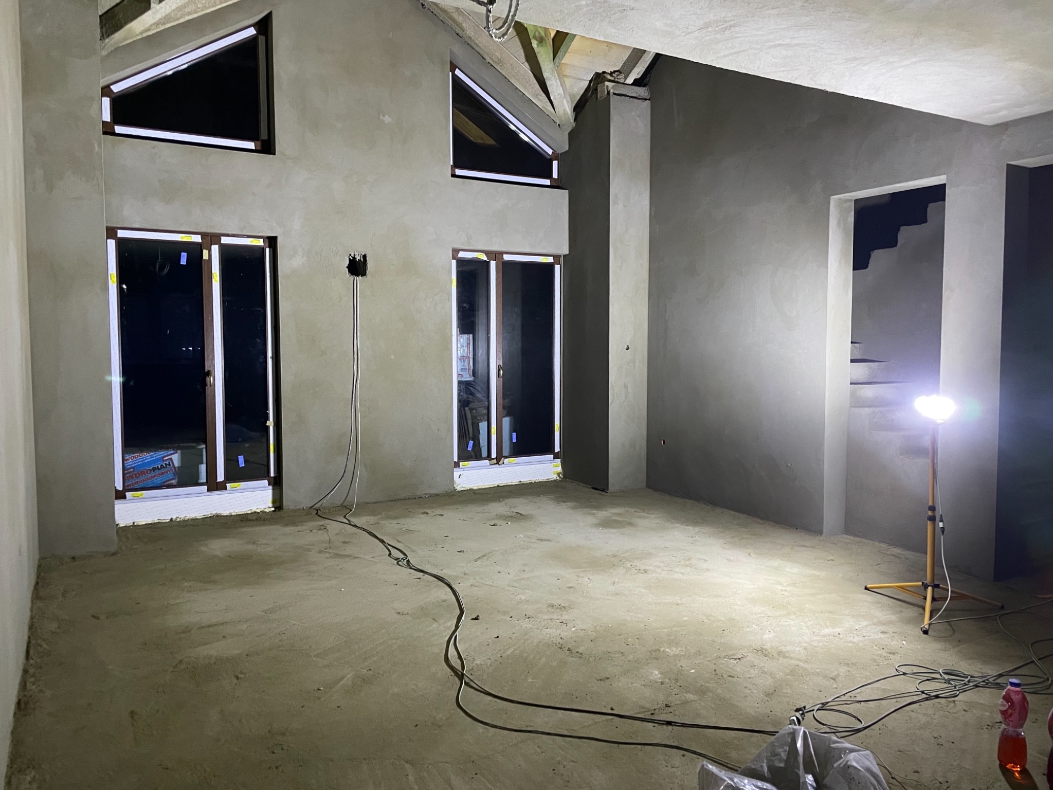 Wnętrze w stanie surowym zamkniętym z dwoma oknami balkonowymi i oknami trójkątnymi pod dachem, ściany pokryte szarym tynkiem, na podłodze wylewka, widoczny reflektor budowlany i kable.