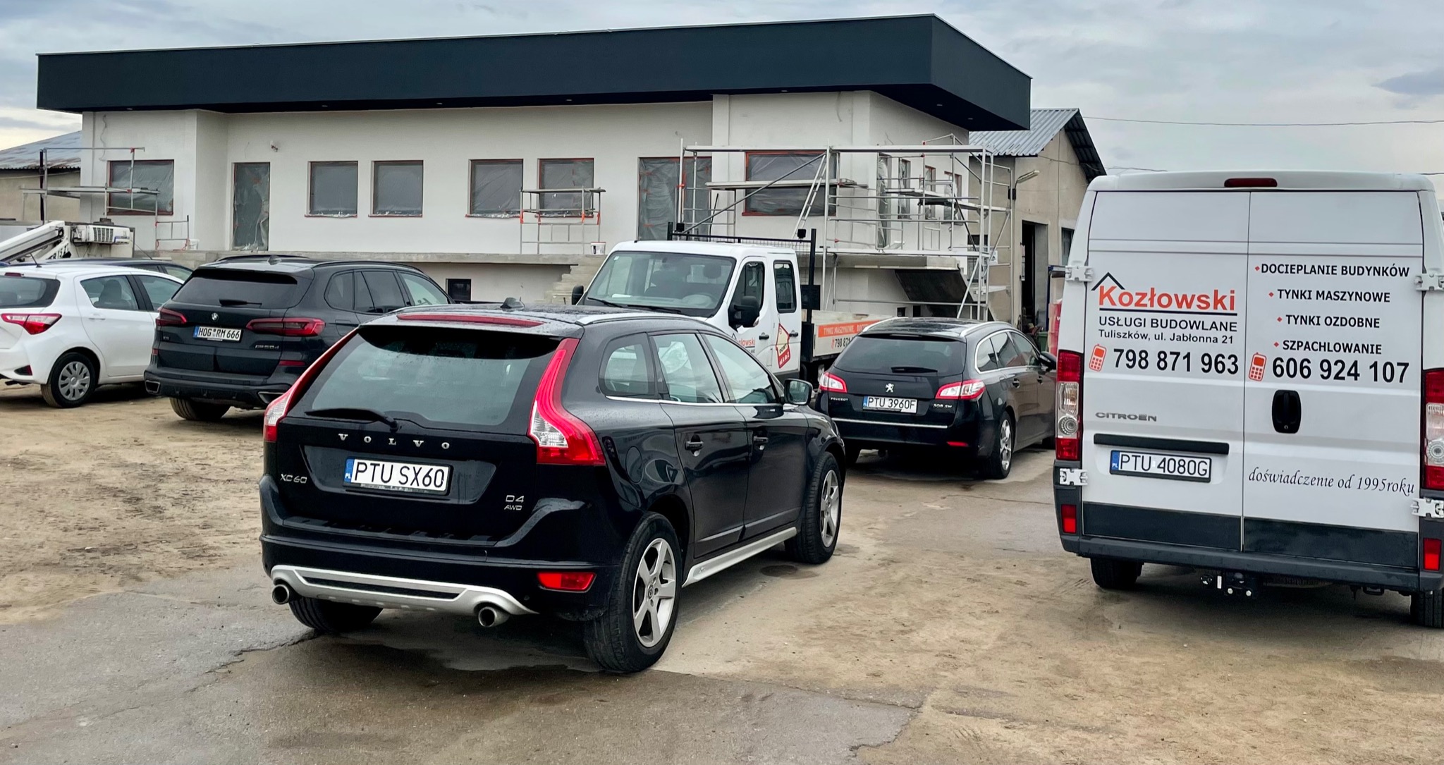 Budynek w trakcie prac elewacyjnych, widoczne rusztowania, zaparkowane samochody osobowe i bus firmy budowlanej z reklamą usług: docieplenia budynków, tynki maszynowe, szpachlowanie.