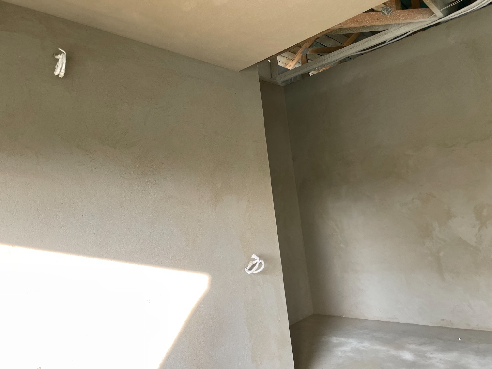 Surowe wnętrze w trakcie wykańczania: świeżo nałożona, jasnoszara zaprawa na ścianach i suficie, widoczne kable elektryczne wystające ze ściany, fragment niezakrytej konstrukcji stropu.