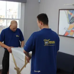 Dwóch mężczyzn w niebieskich koszulkach z logo firmy transportowej zabezpiecza obraz folią bąbelkową i taśmą pakową w jasnym pomieszczeniu z oknem i obrazem na ścianie.