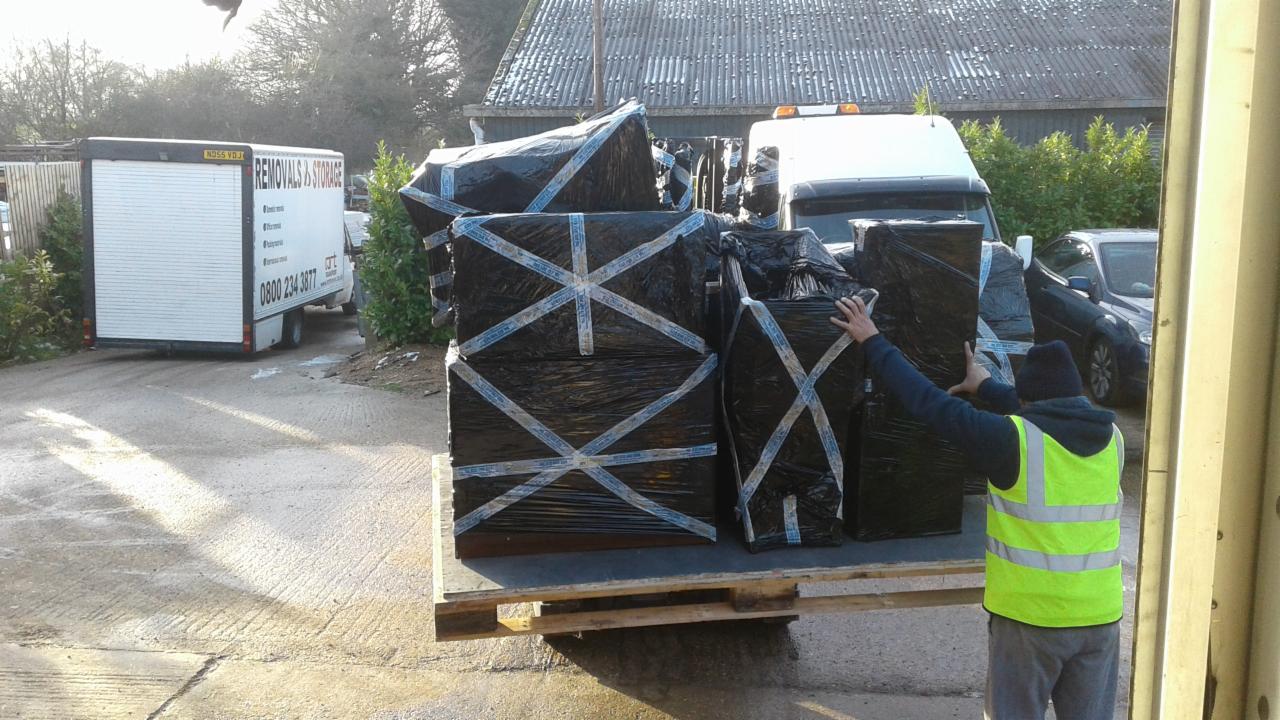 Pracownik w kamizelce odblaskowej zabezpiecza ładunek mebli owiniętych folią i taśmą na palecie wewnątrz kontenera, z widocznym pojazdem firmy przeprowadzkowej w tle.