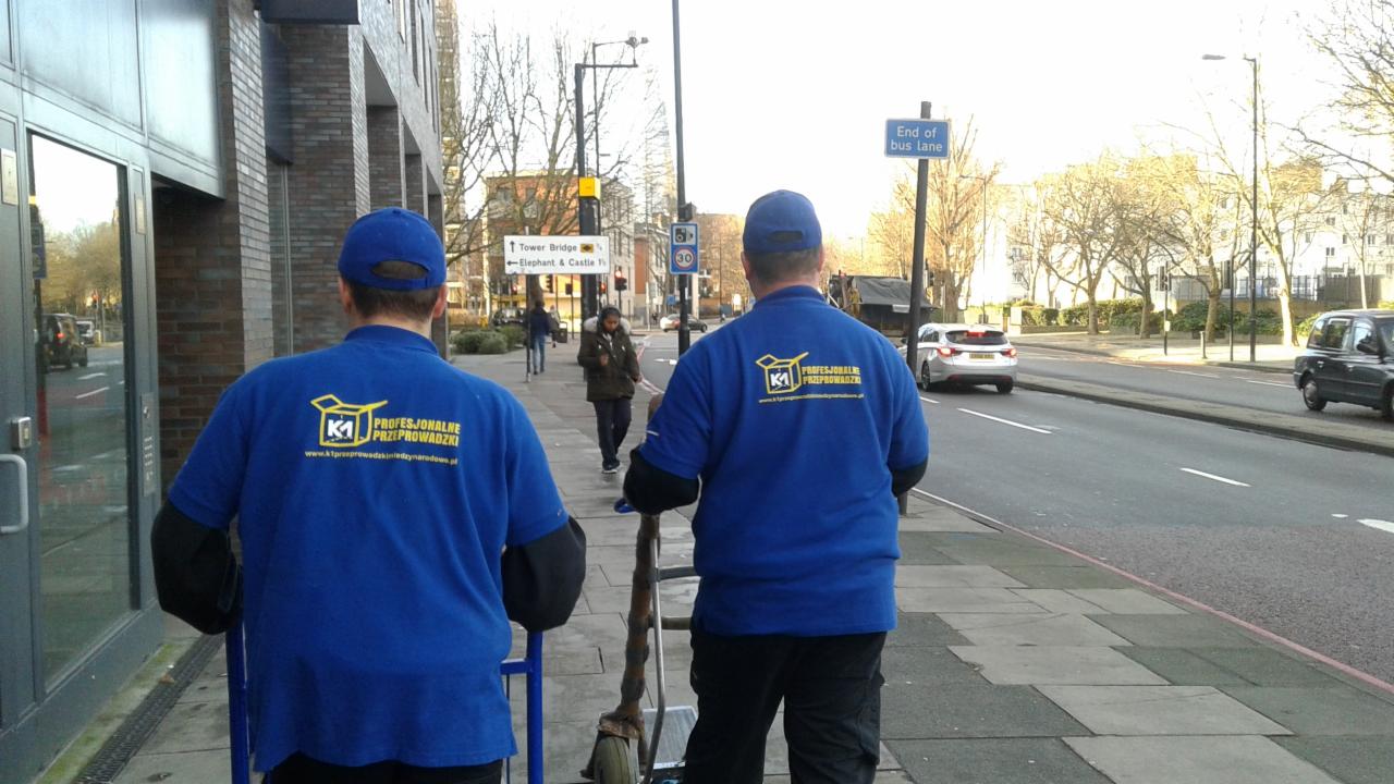 Dwóch mężczyzn w niebieskich uniformach z logo firmy transportowej idących chodnikiem, w tle ulica z ruchem samochodowym i znaki drogowe wskazujące kierunek na Tower Bridge i Elephant & Castle.