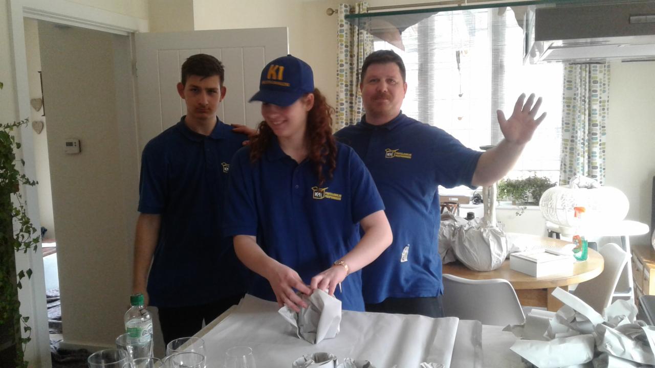 Trzech pracowników firmy przeprowadzkowej w niebieskich uniformach pakuje przedmioty do transportu, używając papieru pakowego, w tle widoczne elementy wyposażenia mieszkania.