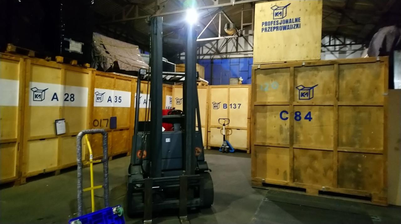 Wnętrze magazynu z drewnianymi skrzyniami oznaczonymi numerami, w tle wózek widłowy i wózek transportowy.