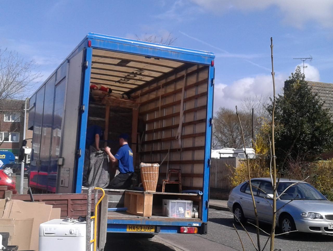 Dwóch mężczyzn w niebieskich koszulkach ładuje zapakowane przedmioty do otwartej ciężarówki z niebieską plandeką, na zewnątrz widoczne kartony, pralka, kosz wiklinowy i srebrny samochód.