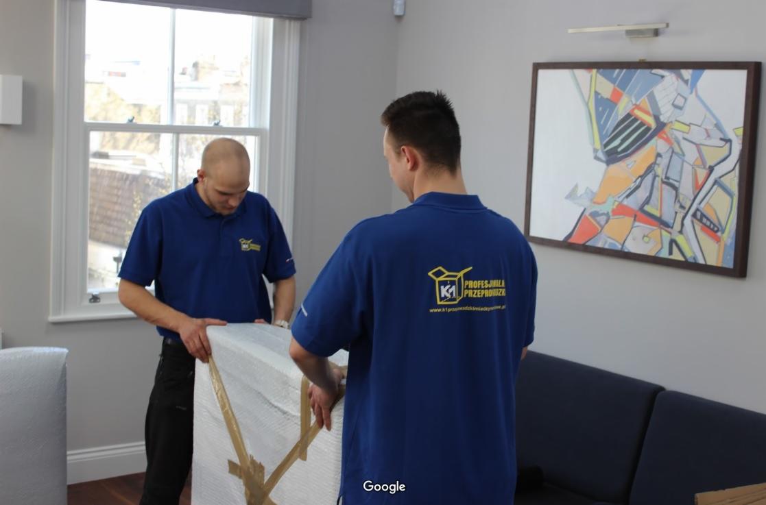 Dwóch mężczyzn w niebieskich koszulkach z logo firmy transportowej zabezpiecza obraz folią bąbelkową i taśmą pakową w jasnym pomieszczeniu z oknem i obrazem na ścianie.