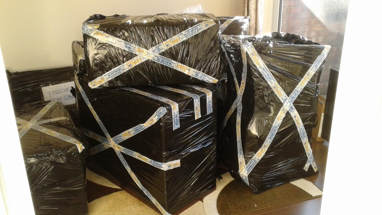 Spakowane meble, owinięte czarną folią i zabezpieczone taśmą z nadrukowanym numerem telefonu, gotowe do transportu; na jednym z pakunków widoczna naklejka z napisem 'Łóżeczko Dom Gdynia'.