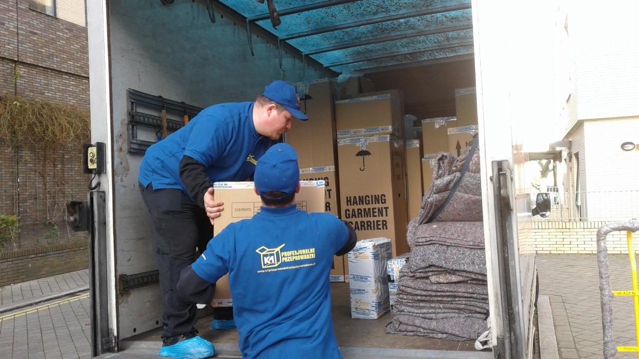 Dwóch pracowników w niebieskich uniformach i ochraniaczach na buty przenosi karton z napisem 'Hanging Garment Carrier' wewnątrz furgonetki wypełnionej kartonami i kocami zabezpieczającymi.