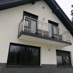 Nowoczesny dom z białymi ścianami, szarą podmurówką z płytek imitujących kamień i balkonem z balustradą ze stali nierdzewnej, okna z roletami zewnętrznymi.