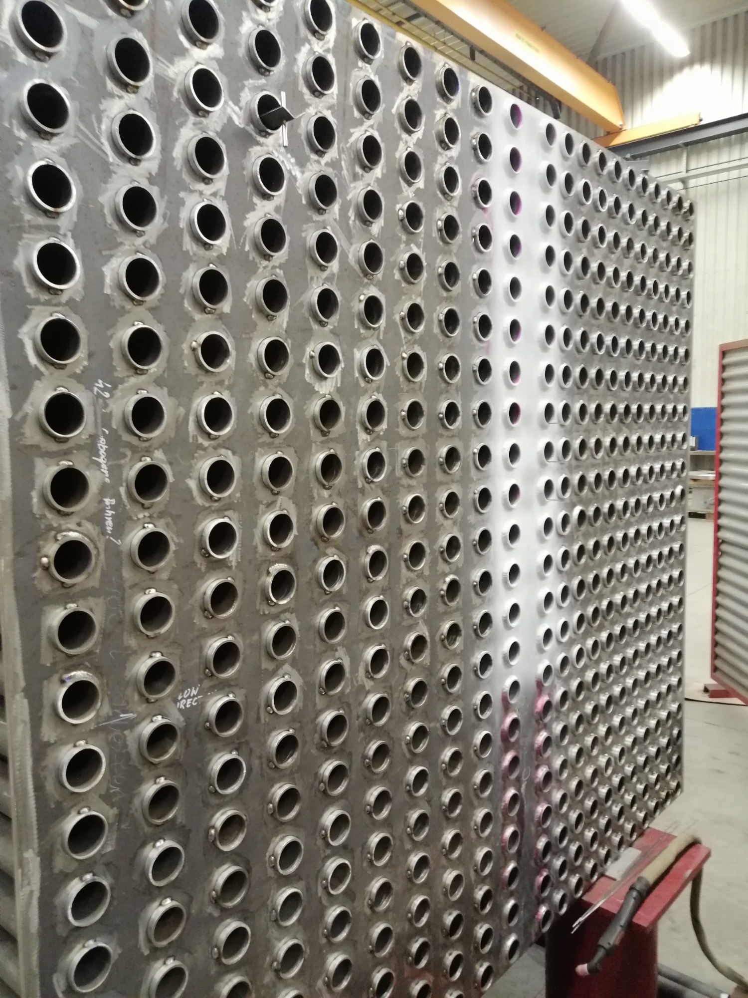 Duży, metalowy wymiennik ciepła z licznymi otworami, częściowo wyszlifowany, widoczny sprzęt spawalniczy i suwnica w tle.