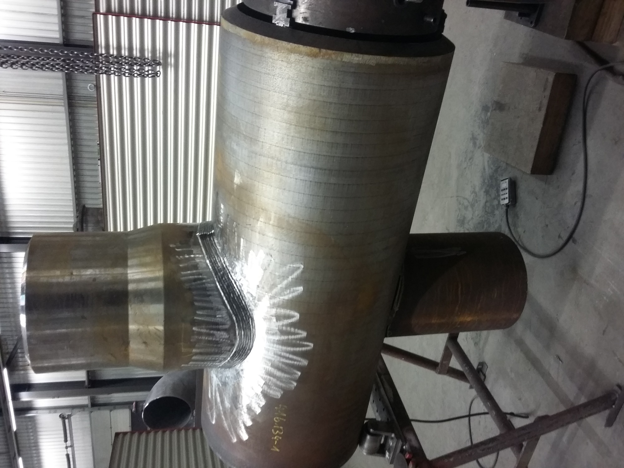 Konstrukcja spawana z rur stalowych na metalowym stojaku, widoczne ślady spawania i numer seryjny na jednej z rur, w tle blaszana ściana i łańcuch.