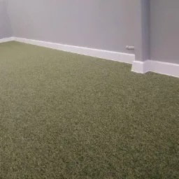 Pokrycie podłogowe z zielonej wykładziny dywanowej, widok na fragment ściany z białą listwą przypodłogową i rurą wystającą ze ściany.