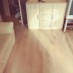 Jasna, drewnopodobna podłoga z widocznymi słojami, częściowo zakryta przez beżową sofę i szafkę z szufladami, oświetlona naturalnym światłem.