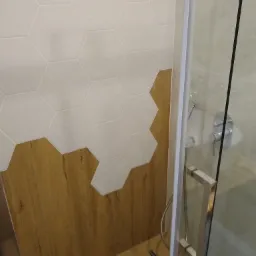 Wykończona ściana w łazience z białymi, sześciokątnymi płytkami przechodzącymi w drewnopodobny panel, widoczny fragment kabiny prysznicowej z chromowaną armaturą.