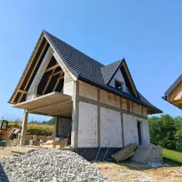 Wznoszenie domu z białych bloczk&oacute;w: widoczna konstrukcja dachu z czarną dach&oacute;wką, betonowy strop nad garażem, wzmocnienia pionowe. Na pierwszym planie hałda gruzu.