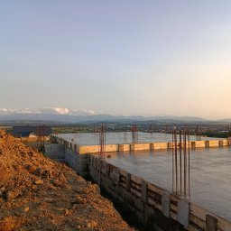 Wojciech Wróbel - Zalany fundament płyty budynku w trakcie budowy, z wystającymi prętami zbrojeniowymi. W tle widok na panoramę miasta i góry w oddali, oświetlone zachodzącym słońcem.