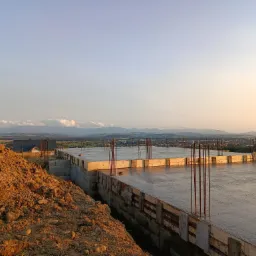 Zalany fundament płyty budynku w trakcie budowy, z wystającymi prętami zbrojeniowymi. W tle widok na panoramę miasta i góry w oddali, oświetlone zachodzącym słońcem.